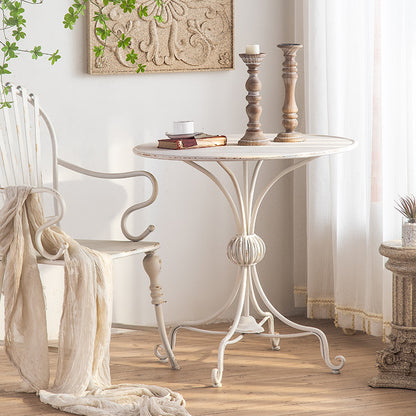 A rustic, round Unwind in Style: The White American Balcony Table by Furnishaus sits on a wooden floor with two brown candlesticks and an open book. An ornate white chair with draped fabric complements the vintage ambiance of white curtains and a textured wall decoration. ShopFurnishaus.com achieves The Enchantment.