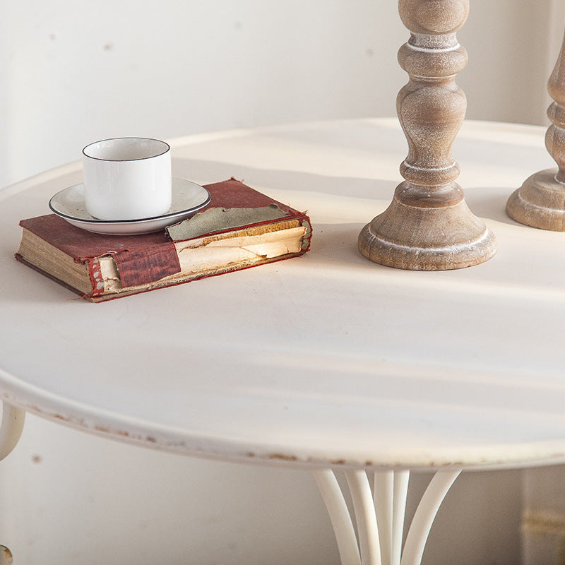 In the soft sunlight, Unwind in Style: The White American Balcony Table by Furnishaus - Home and Living hosts a weathered book with a red cover and a white teacup on a saucer. Two light brown wooden candlesticks accompany the scene, evoking vintage charm reminiscent of ShopFurnishaus.com.