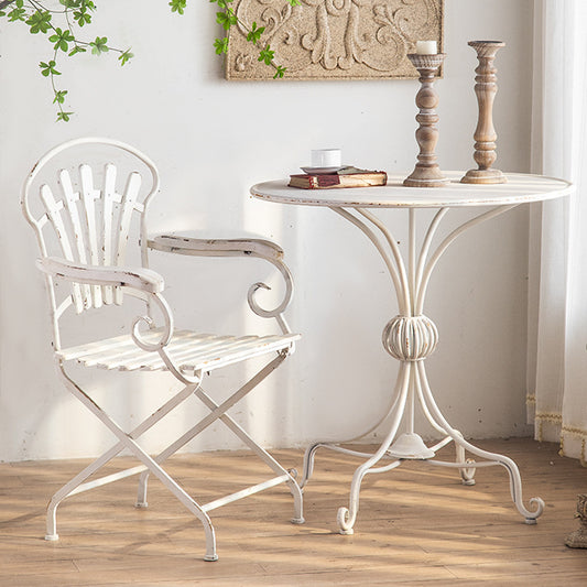 A rustic white metal chair with ornate armrests complements the Unwind in Style: The White American Balcony Table by Furnishaus - Home and Living. Two candles in wooden holders add charm, while a book and cup sit gracefully atop. Set on a wooden floor, green leaves and a decorative wall panel complete the scene.