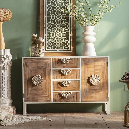 The Modern Farmhouse Sideboard Cabinet with Intricate Carvings by Furnishaus - Home and Living adds rustic charm against a mint green wall. A geometric-patterned mirror hangs above, while a large white vase of blossoming branches and two smaller decorative vases adorn the top.