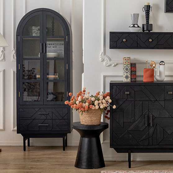 A room with ornate white wall paneling showcases modern design, featuring Furnishauss Arched Glass Door Display Cabinet with Geometric Accents on the left, displaying decor. A black side table holds vibrant flowers beside it, and a black sideboard with geometric patterns sits on the right.