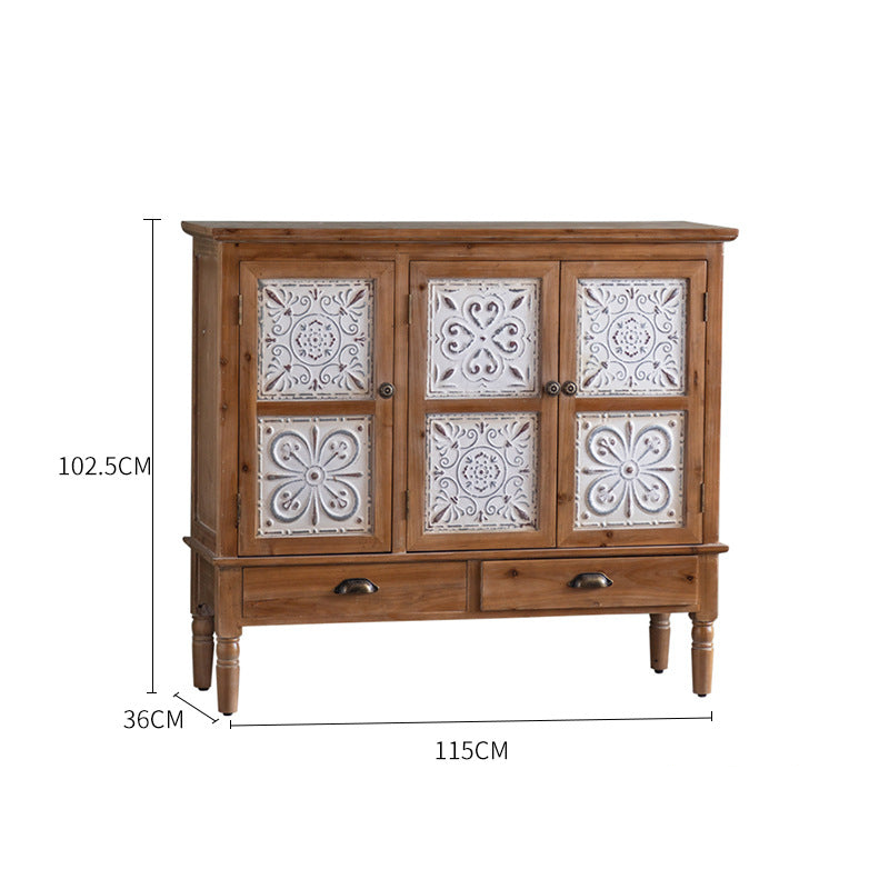 The Rustic Sideboard by Furnishaus boasts a timeless design with decorative white carved floral panels on its two doors, a measurement of 102.5cm tall, 115cm wide, and 36cm deep. It includes two drawers and stands on four legs with metal knobs, highlighting quality craftsmanship and rustic charm.